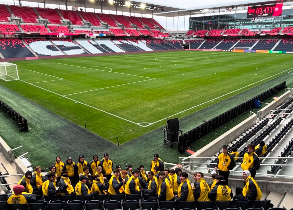 Lewis and Clark students in yellow jackets sitting in the stands of a large, empty soccer stadium overlooking the field that has the word "CITY" on the other side of the stands.