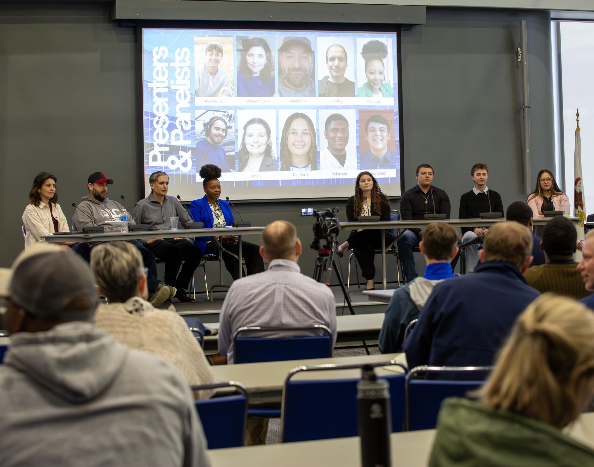 A panel of speakers sits at a table in front of a screen displaying a grid of their headshots.
