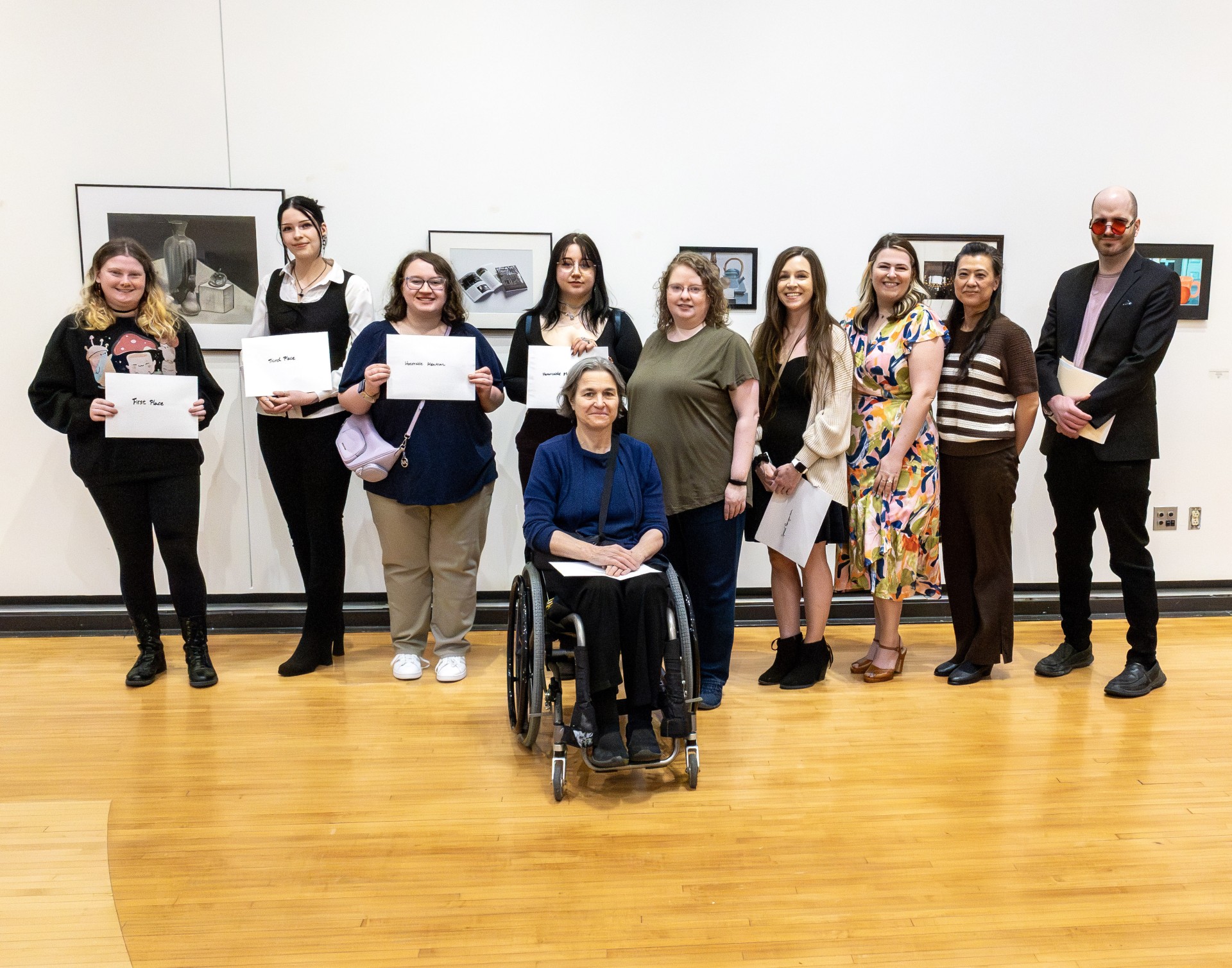 A group photograph taken inside an art gallery with light wooden floors, showing ten individuals posing together in front of a white wall displaying framed artwork. Four students on the left hold white sheets of paper indicating contest placements, while a woman in a blue shirt sits in a wheelchair at the center of the group.