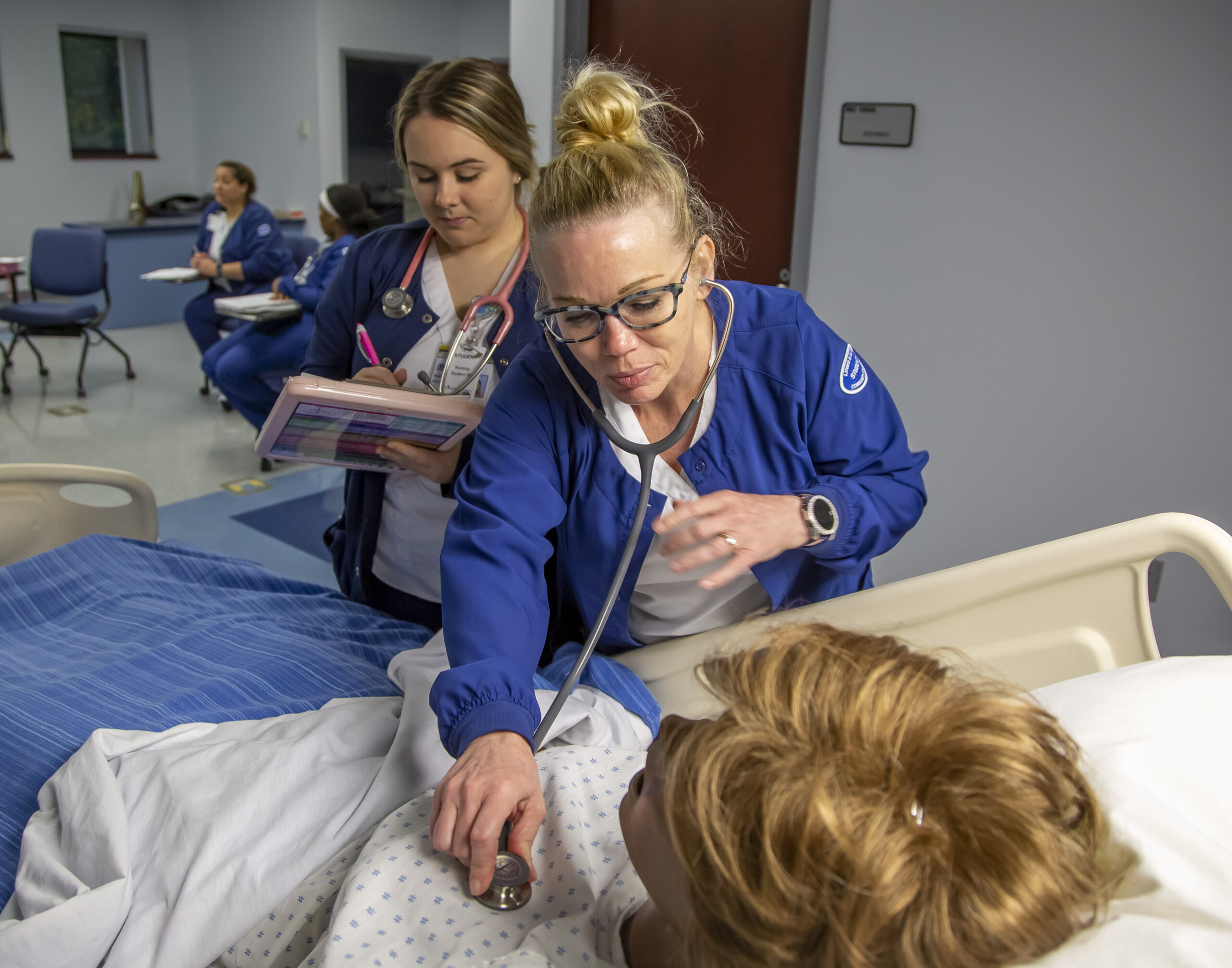 A nursing student uses a stethoscope to examine a dummy patient in a medical training room while another student takes notes nearby.