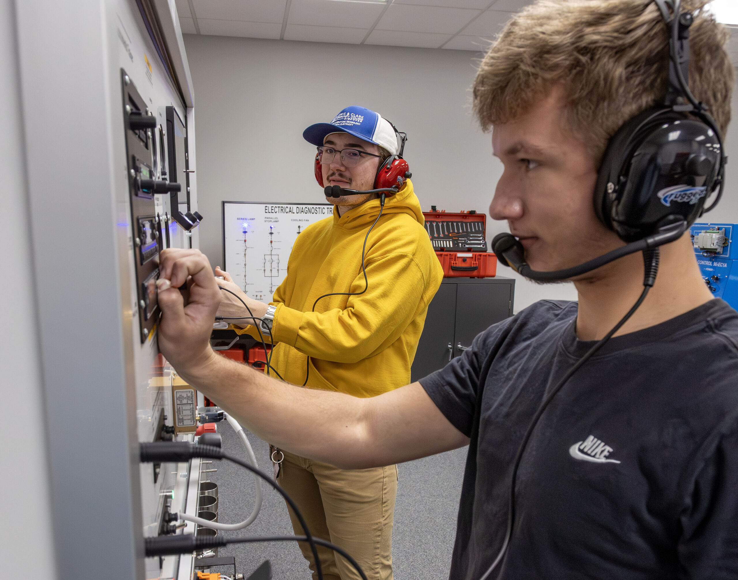 Two students wearing headsets work together on an electrical diagnostic training board.