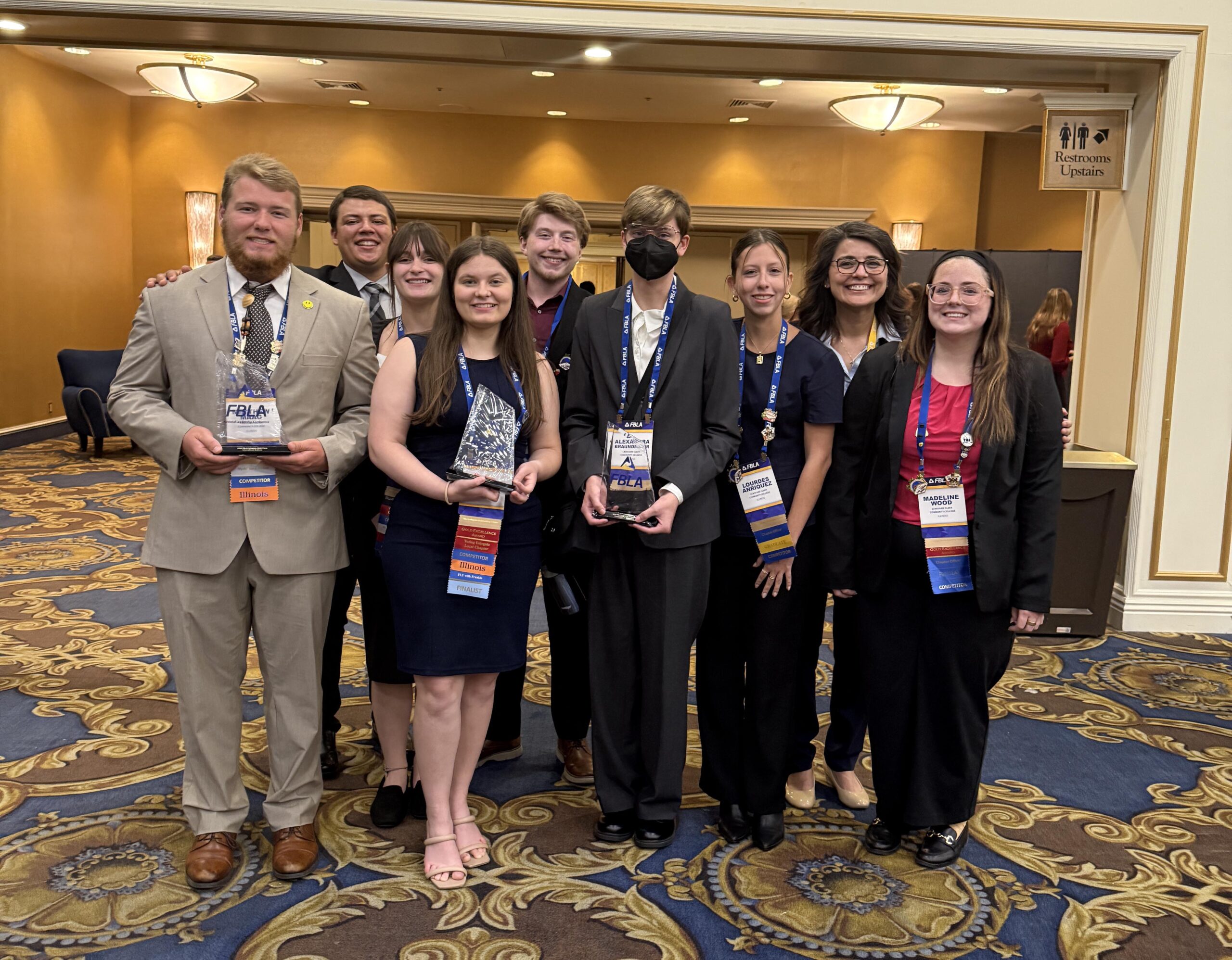 A group of FBLA students and a staff member stand together smiling in a hotel hallway, some holding awards and wearing competition lanyards.