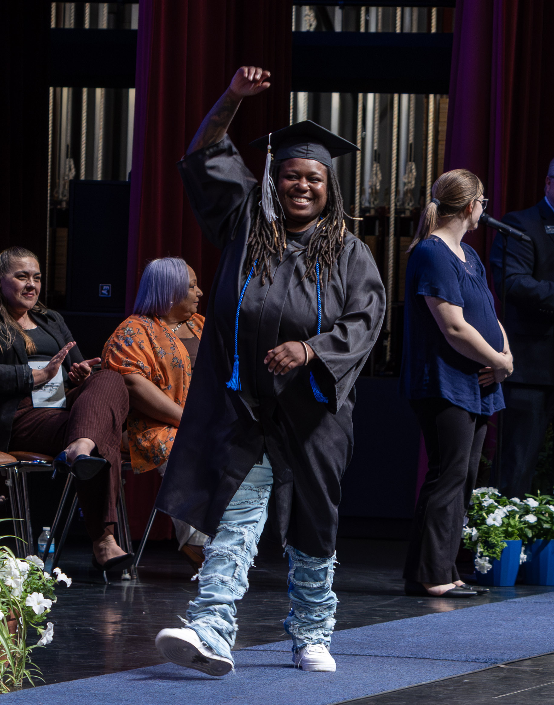 A smiling graduate in a cap and gown with one arm raised in celebration, walking across a stage.