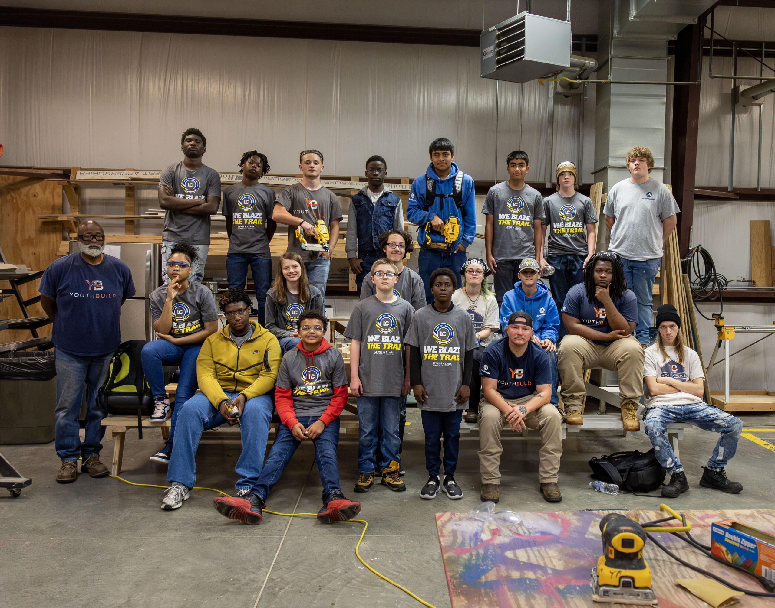 A group of YouthBuild students and instructors pose together in a workshop space, many wearing 'We Blaze the Trail' T-shirts.