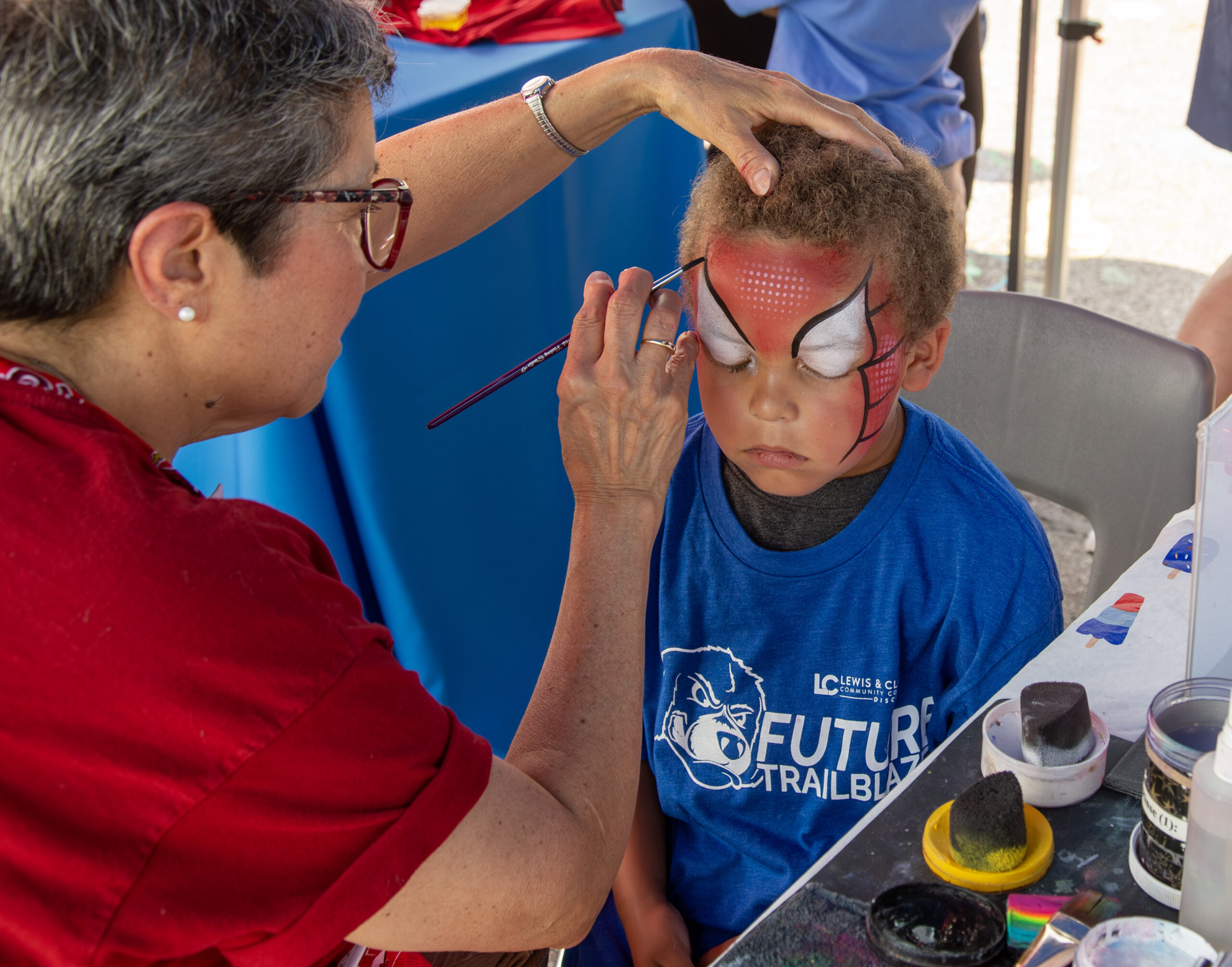 A face painter uses a fine brush to carefully apply a red and white Spider-Man mask design to a young boy's face at an outdoor community event.