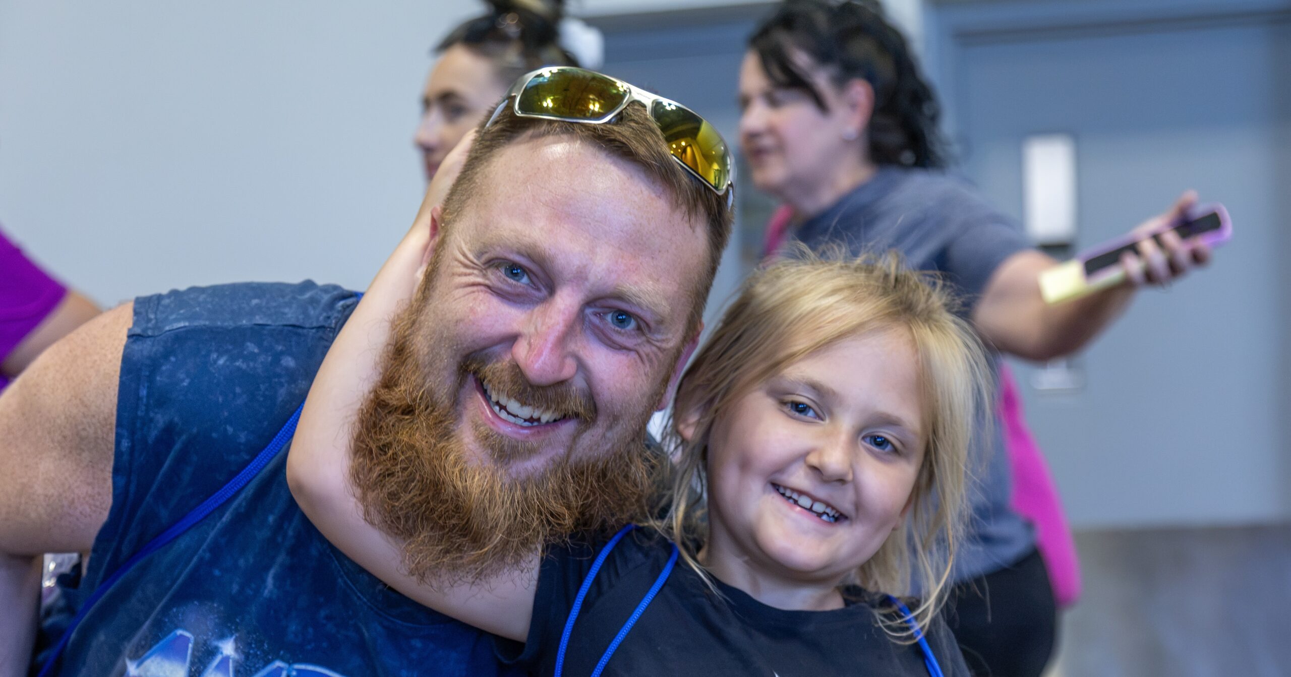 A bearded man and a young girl smile while embracing, with two women visible in the background.