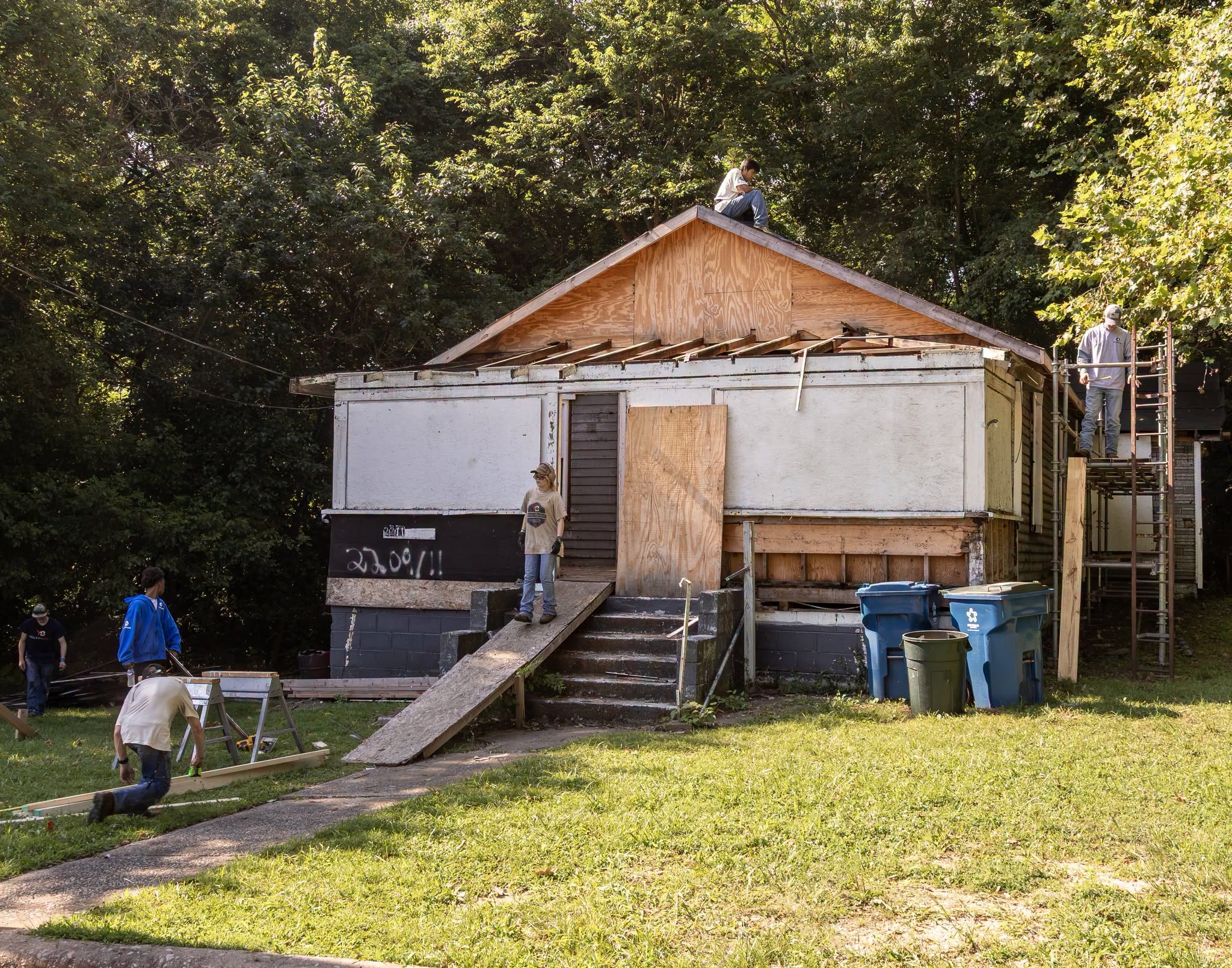 A group of people working on the exterior renovation and construction of a house surrounded by trees and a grassy lawn.