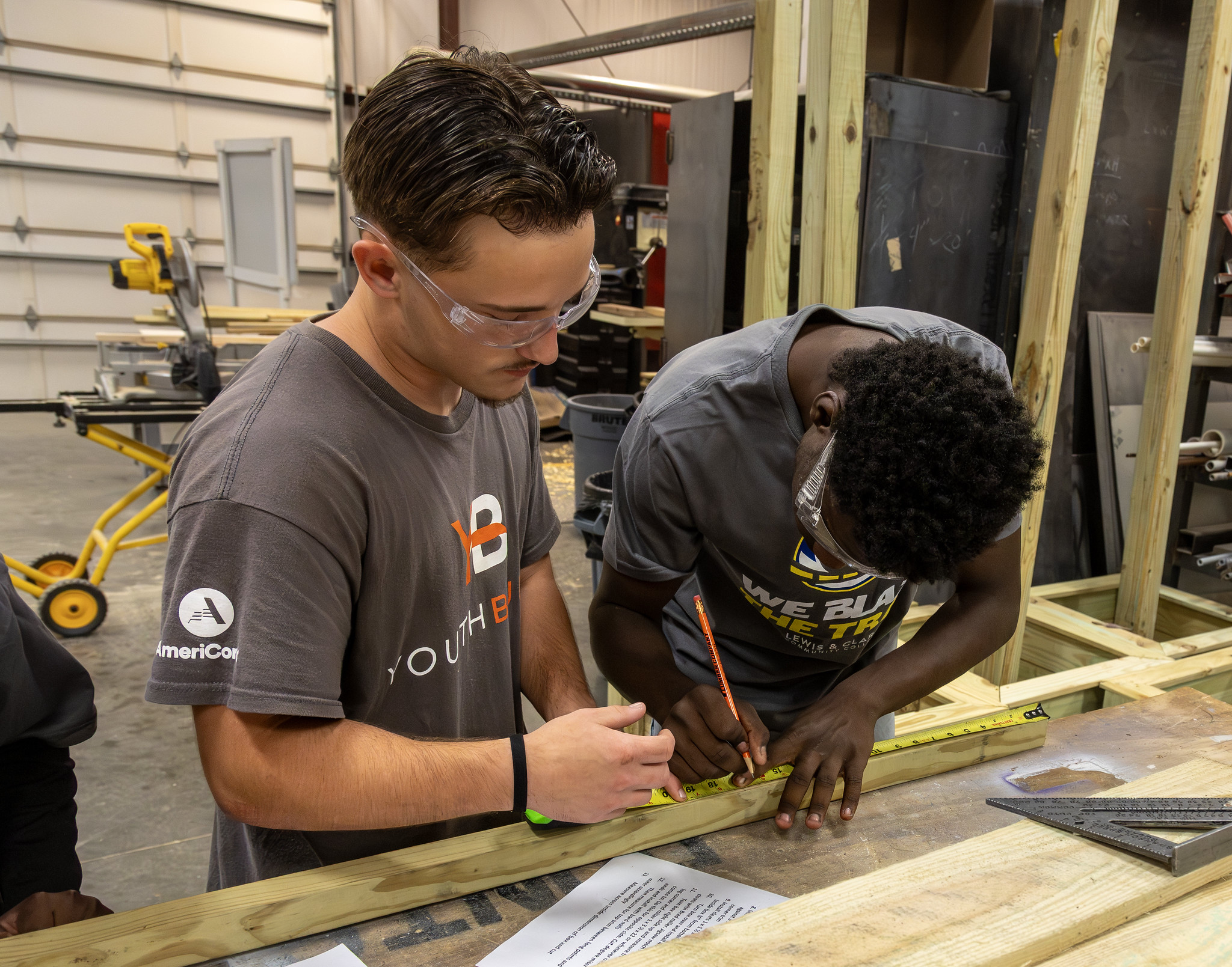Two students in a workshop measure a wooden board with a tape measure and mark it with a pencil.