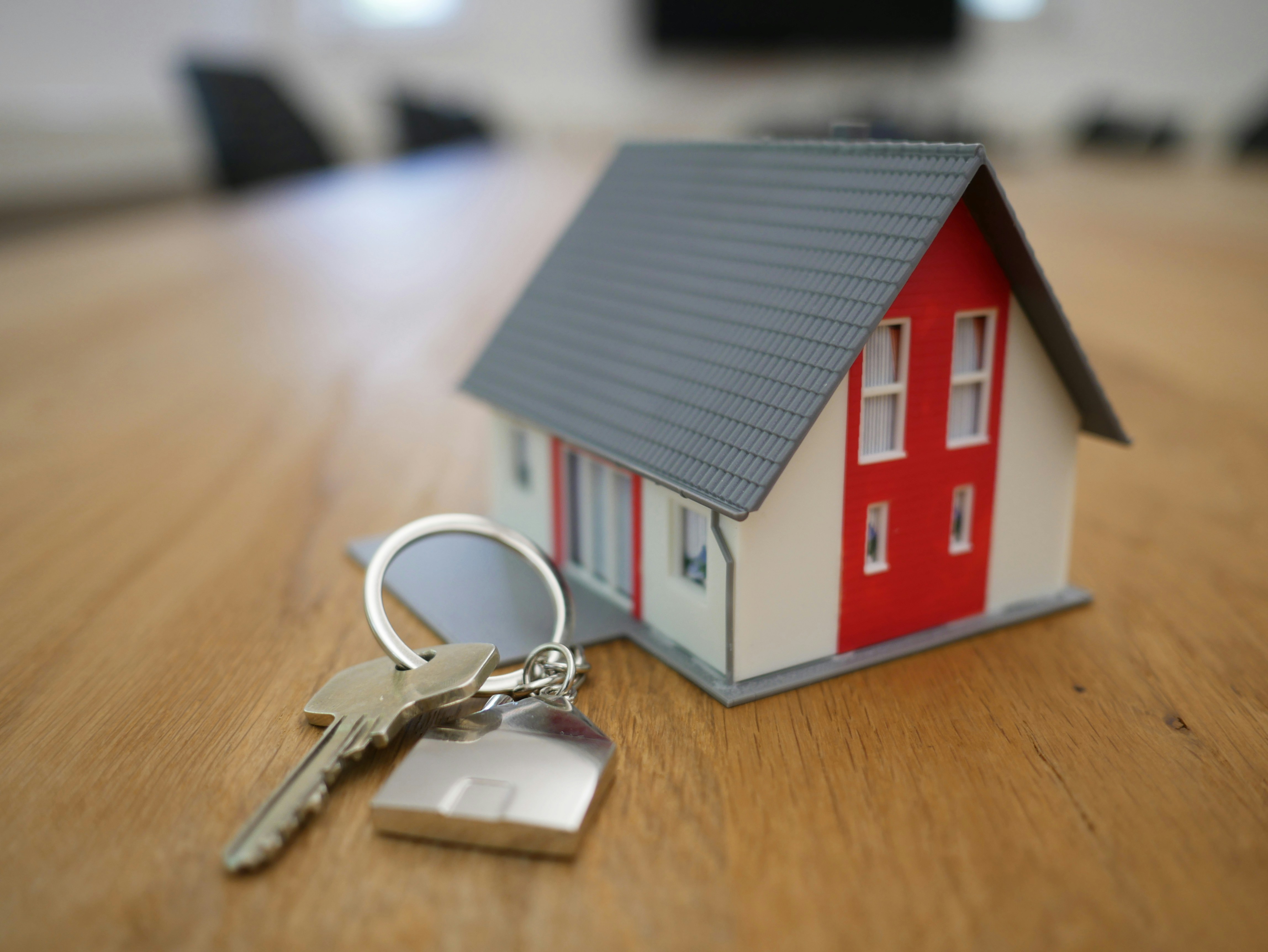A model house with a grey roof and red trim sitting on a wooden table next to a keychain with keys and a house-shaped charm.